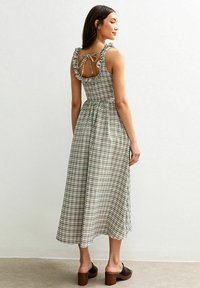 Vrouw in een mouwloze, groen-wit geruite midi-jurk met ruffles op de schouders, vastgebonden in een strik aan de achterkant, staand tegen een witte muur.
