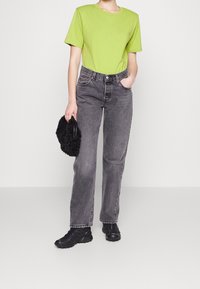 Camiseta verde de manga corta, pantalones vaqueros negros de cintura alta, bolso texturizado negro y zapatillas deportivas negras, de pie sobre un fondo neutro.