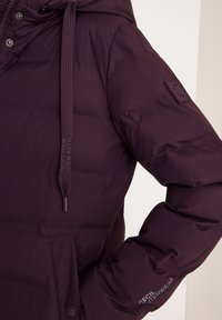 Giacca invernale imbottita viola con cappuccio, cordini, una tasca sul petto e il testo "CECIL OUTERWEAR" sulla manica.