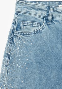 Falda de mezclilla azul claro con un bolsillo, adornada con tachuelas plateadas dispersas para añadir textura y detalle.