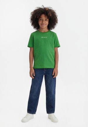 Dijete s kovrčavom kosom u zelenoj majici "LES DEUX", plavim trapericama i bijelim tenisicama, stoji na jednobijeloj pozadini.