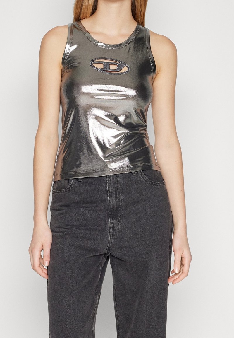 Femme portant un débardeur métallique argenté sans manches avec un logo découpé ovale sur la poitrine et un jean noir taille haute.