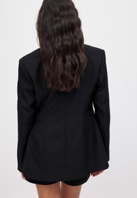 Juodas pritaikytas švarkas, pagamintas iš tekstūruoto audinio, turintis prigludusią siluetą, struktūruotus pečius ir platų apdailą, matomas iš nugaros.