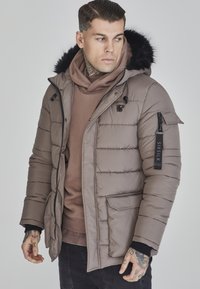 Polstret brun yderjakke med hætte foret med pels, lynlåslomme på ærmet, tekstureret stof og lagdelt med en brun sweater.