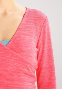 Tričko s dlouhým rukávem a výstřihem do V v jasně růžové barvě, vyrobené z měkké, mírně strukturální tkaniny s překládaným designem. Převlečené přes světle modrou vrstvu.