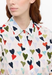 Camicia bianca con colletto caratterizzata da un motivo di cuori colorati in rosso, verde, blu e nero. Include bottoni bianchi. Tessuto morbido.