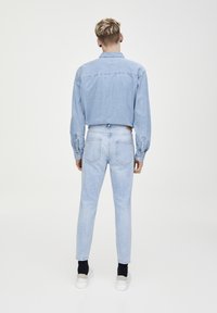 Camisa de mezclilla azul claro y jeans, diseño ajustado, con puños abotonados y bolsillos traseros. Modelo lleva zapatillas blancas y calcetines negros.
