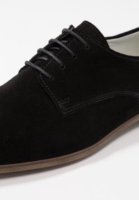 Scarpa oxford in suede nero con sei occhielli, lacci piatti neri, dettagli di cucitura discreti e suola in gomma marrone. Texture liscia.
