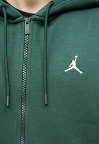 Felpa verde con zip realizzata in tessuto morbido, caratterizzata da un logo Jumpman bianco ben visibile. Include lacci e una zip testurizzata.
