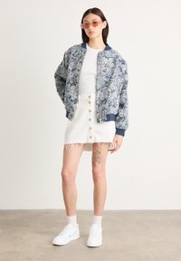 Veste bomber fleurie bleue, débardeur blanc, mini-jupe en jean blanche avec boutons, baskets blanches et lunettes de soleil roses. Tatouage apparent sur la jambe.