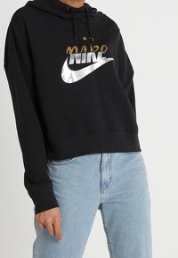 Person som bär en svart Nike-hoodie med metallisk logotyp och ljusblå jeans, stående mot en helt vit bakgrund.