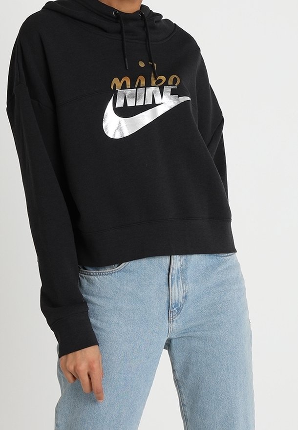 Person som bär en svart Nike-hoodie med metallisk logotyp och ljusblå jeans, stående mot en helt vit bakgrund.