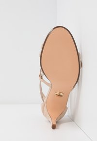 Beige högklackad sko med spetsig tå, mockaliknande yta och en slät gummisula med räfflor och en metallisk logotypdetalj.