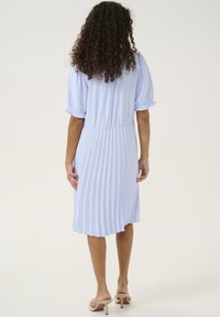 Nabrano svetlo modra obleka z kratkimi, napihnjenimi rokavi, elastičnim pasom in fluidno zadnjo stranjo. Nosijo se s sandalami na peto v nevtralnih barvah.