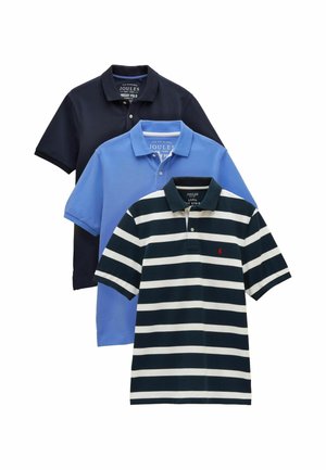 Man met gevouwen armen, gekleed in een lichtblauw poloshirt met korte mouwen en beige broek, staand tegen een effen beige achtergrond.