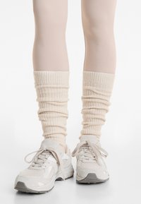 Hvide strikkede benvarmere med ribbet tekstur, parret med lysegrå sneakers, der har en kombination af ruskind og mesh-materialer, rund tå og snørebånd.