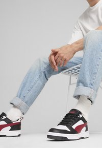 Černobílé tenisky s červenými akcenty, vyrobené z kůže a gumy. Čistý design s tkaničkami, nošené s světle modrými džíny a bílými ponožkami.