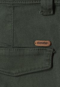 Dunkelgrüner Stoff mit einer genähten Tasche. Ein kleines braunes Lederetikett mit dem Text "minymo" ist befestigt und zeigt die Markeninformationen.