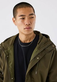 Olijfgroene hooded jas met een rits en trekkoorden, gelaagd over een zwart shirt, met zilveren accenten en een gladde textuur.