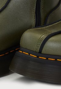 Gros plan sur des bottes en cuir vert olive avec semelles noires et détails de couture orange.