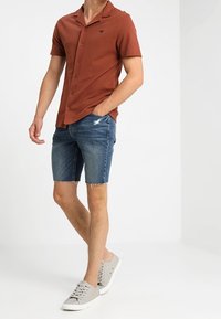 Hnědá košile s krátkým rukávem na knoflíky, kombinována s džínovými šortkami s třepenými okraji a světle šedými teniskami s tkaničkami.