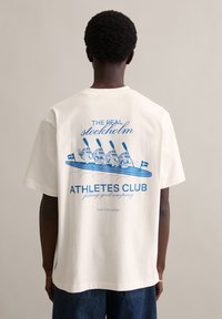 Witte katoenen T-shirt met een blauwe grafische print van cartoon-peddelaars in een kano, met de tekst 'THE REAL STOCKHOLM ATHLETES CLUB.'