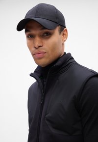 Colete preto sem mangas com forro de fleece e tecido liso, apresentando um fecho de correr na frente e um logotipo no peito. Boné preto complementa o visual.