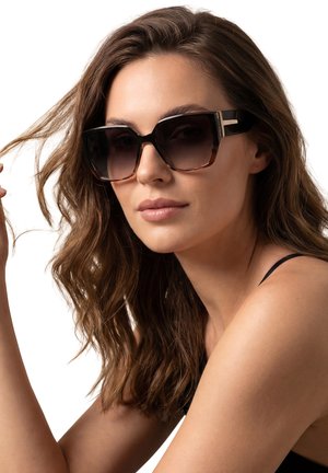 Kobieta o falowanych brązowych włosach nosząca duże kwadratowe brązowe okulary przeciwsłoneczne, trzymająca kosmyk włosów, na białym tle.