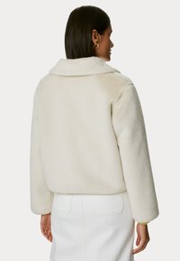 Límcová, krémová bunda z umělé kožešiny s oversized střihem, lehce zkrácenou délkou a texturovaným povrchem. Nošena přes bílou sukni.