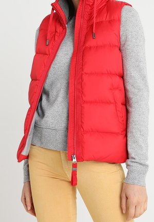 Rode gewatteerde mouwloze bodywarmer over een grijze trui met lange mouwen en beige broek, met de bodywarmer gedeeltelijk opengezipped.