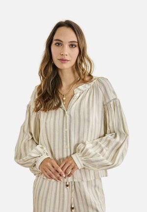 Mujer con cabello largo y ondulado que lleva una blusa beige a rayas con botones y mangas abullonadas, combinada con pantalones a juego y un collar dorado.