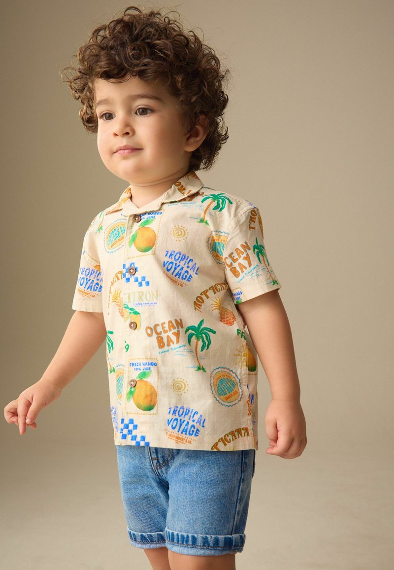 Niño pequeño con cabello rizado que lleva una camisa beige con estampado tropical y pantalones cortos de mezclilla remangados, de pie frente a un fondo sencillo.