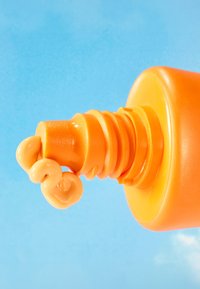 Botella exprimidora naranja con un dispensador texturizado, emitiendo un producto suave, cremoso y de color naranja claro sobre un fondo azul claro.