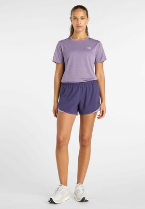 Vrouw staand, gekleed in een lichtpaars sportshirt met korte mouwen, donkerpaarse hardloopshorts en witte hardloopschoenen, tegen een witte achtergrond.