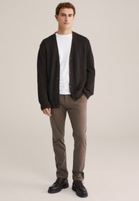 Cardigan tricotat de culoare neagră, cu decolteu în V, închidere cu buton, purtat peste un tricou alb, asortat cu pantaloni maro și încălțăminte neagră cu șireturi.