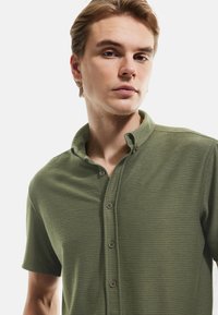 Camisa de botones de manga corta en verde oliva con textura acanalada, un cuello y cinco botones marrones en el frente.