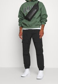 Zielona bluza z kapturem z białym napisem, czarne spodnie bojówki z kieszeniami oraz białe trampki. Torba jest czarna, teksturowana i noszona przez ramię.