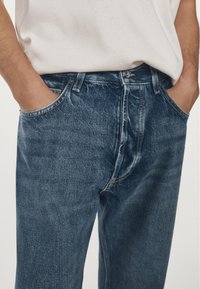 Jeans in denim blu scuro con vestibilità rilassata, dotati di tasche frontali, chiusura con bottoni e dettagli di cuciture visibili. Tessuto strutturato.