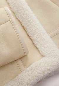Beige schapenvacht jackdetail met zachte, pluizige voering en een gladde buitenkant. Heeft gestikte randen en een subtiele glans.