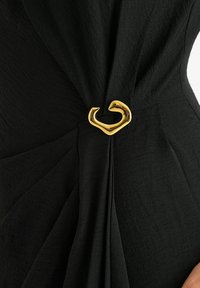 Black fabric garment with intricate pleating and a gold, curved metal accent. The texture is soft with a slight sheen.