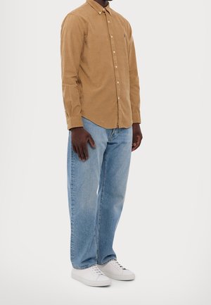 Mand iført en brun kordfløjls-skjorte med knapper, lyseblå jeans og hvide sneakers, stående mod en ensfarvet baggrund.