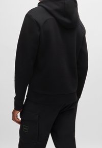 Felpa nera con cappuccio, tasca a marsupio, polsini e orlo a coste. Presenta un cappuccio e dettagli di cucitura visibili. Abbinata a pantaloni jogger neri coordinati.