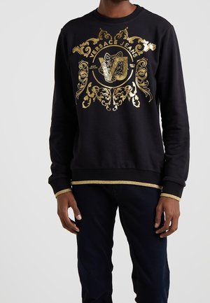 Sweat-shirt noir à manches longues avec un imprimé doré orné représentant une tête de tigre et le texte « Versace Jeans », porté par une personne debout.