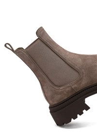 Bottine Chelsea en daim marron avec des panneaux latéraux élastiques, une languette et une semelle en caoutchouc robuste avec des crampons profonds. Texture lisse et forme arrondie.
