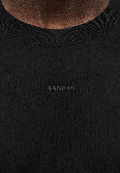 Κοντινό πλάνο ενός ατόμου που φοράει μαυρο φούτερ με στρογγυλή λαιμόκοψη, με τη λέξη "SANDRO" τυπωμένη με μικρά, διακριτικά μαύρα γράμματα στο στήθος.