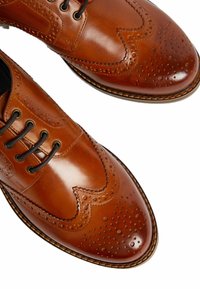 Zapatos de vestir de cuero marrón con un diseño tipo wingtip, detalles perforados y costuras en contraste a lo largo de la suela; cierre con cordones.