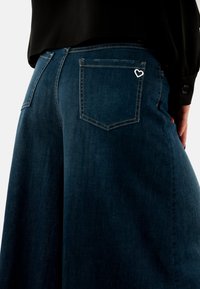 Pantalones de mezclilla de pierna ancha en azul marino con un acento plateado en forma de corazón en el bolsillo trasero, que presentan costuras clásicas y una textura suave.