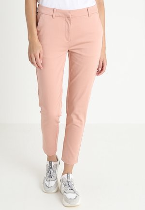 Vrouw draagt slanke, enkellange lichtroze broek gecombineerd met witte en grijze sneakers, staand op een witte achtergrond.