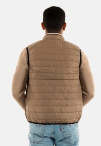 Gilet rembourré marron avec des coutures horizontales, col montant et finitions noires ; porté sur un pull beige et associé à un jean bleu clair.