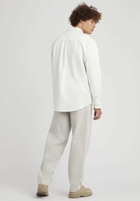Chemise blanche à col boutonné avec un ourlet arrondi et un détail au dos, associée à un pantalon gris clair à coupe ample et des chaussures beiges.